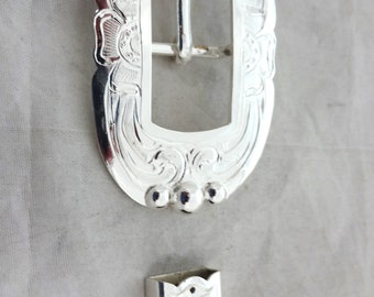 May include: Silver belt buckle with an ornate design and a matching tip. The buckle has a rectangular opening for the belt and is decorated with a floral pattern.