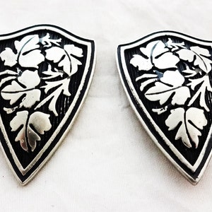 May include: A pair of silver and black shield-shaped earrings with a floral design.