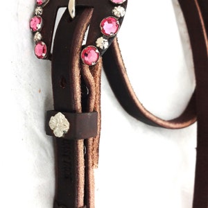 Puede incluir: Un cinturón de cuero marrón con una hebilla plateada decorada con diamantes de imitación rosas. La hebilla tiene un diseño floral.
