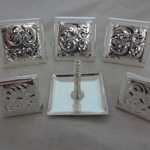 May include: Six silver-toned square-shaped conchos with an intricate floral design. The conchos are arranged in a circle around a single concho with a screw. The screw is silver-toned and has a square head.
