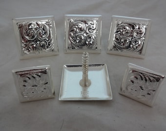 May include: Six silver-toned square-shaped conchos with an intricate floral design. The conchos are arranged in a circle around a single concho with a screw. The screw is silver-toned and has a square head.
