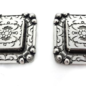Puede incluir: Dos hebillas de cinturón cuadradas plateadas con un intrincado diseño floral.