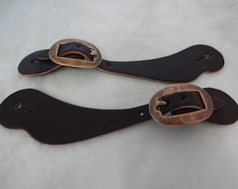 Può includere: Due cinturini per speroni in pelle marrone con fibbie in ottone. I cinturini sono curvi e hanno un foro all'estremità. Le fibbie sono di forma ovale e hanno una finitura liscia.