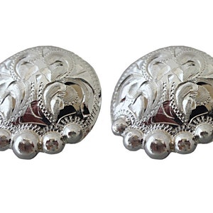 May include: A pair of silver-toned metal conchos with a floral design. The conchos have a rounded shape and a raised edge. They are decorated with a series of small, round beads around the edge.