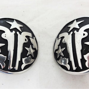 Puede incluir: Un par de pendientes de tono plateado con fondo negro. Los pendientes presentan un diseño de dos pistolas, estrellas y formas florales.