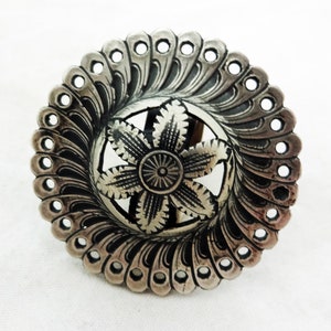 Puede incluir: Un colgante circular de plata con un diseño floral detallado. El colgante tiene un borde festoneado con pequeños agujeros alrededor del perímetro.