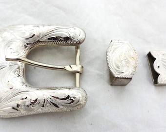 Könnte beinhalten: Ein silberfarbenes Gürtelschnallen-Set mit einem verzierten, gravierten Blumendesign. Das Set enthält die Schnalle, eine Gürtelschlaufe und eine Gürtelspitze. Das Metall hat eine glänzende Oberfläche.