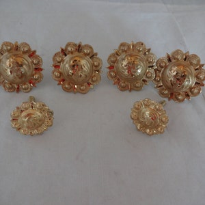 Puede incluir: Un conjunto de botones y tachuelas decorativas de metal dorado. Los botones y tachuelas tienen un diseño floral con un patrón central en relieve. El conjunto incluye seis piezas, con diferentes tamaños y formas, todas con un acabado brillante.