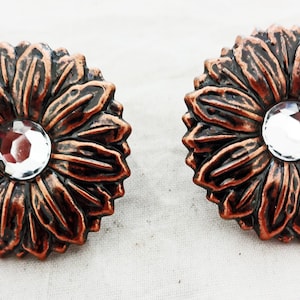 Op de afbeelding: Een paar koperkleurige oorbellen met een bloemmotief en een helder kristal in het midden van elke bloem.