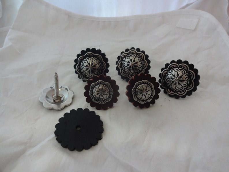 Puede incluir: Conjunto de siete conchos de cuero negro y plateado con un dise&ntilde;o floral decorativo. Los conchos se utilizan para decorar sillas de montar, cinturones y otros art&iacute;culos de cuero.