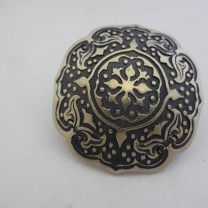 Könnte beinhalten: Eine silberfarbene Brosche mit einem komplizierten schwarzen und weißen Design. Das Design zeigt eine zentrale Blume mit sternförmiger Form, umgeben von kleineren floralen Motiven und Punkten.