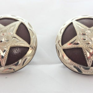 Puede incluir: Un par de conchos de metal plateado con un centro de cuero marrón y un diseño de estrella de cinco puntas.