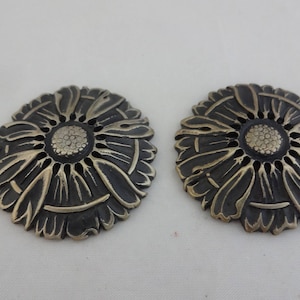 Op de afbeelding: Twee decoratieve metalen bloemversieringen met een zwarte en gouden afwerking. De versieringen zijn rond en hebben een reliëfdesign van een bloem met meerdere bloemblaadjes.