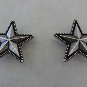 May include: Two silver-toned metal star-shaped embellishments with a beaded border. The stars have a raised, textured surface.