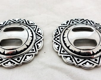 May include: Two silver-toned metal buckles with a decorative design. Each buckle has a circular shape with a center oval cutout. The design features a repeating pattern of triangles and rounded shapes.