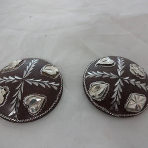May include: Two round, brown, decorative buttons with silver leaf designs. The buttons have a raised edge and a silver leaf pattern in the center.