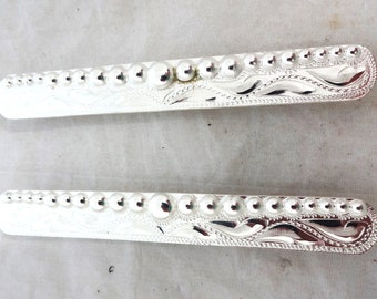 May include: Two silver belt buckles with a decorative design of a stylized floral pattern and a row of raised circles.