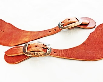 Può includere: Un paio di cinturini per speroni in pelle marrone con fibbie in ottone. I cinturini sono progettati per essere fissati a una sella e servono a tenere gli speroni in posizione.