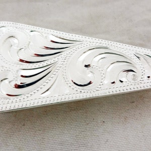 以下が含まれることがあります： 銀色の長方形のベルトバックル。装飾的な彫刻が施された花の模様があります。バックルは丸みを帯びた端と詳細な縁取りがあります。彫刻された模様は、渦巻く葉と蔓を特徴としています。