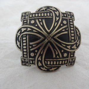Puede incluir: Objeto decorativo cuadrado de metal con un diseño intrincado. Presenta un patrón central en forma de cruz con líneas curvas y detalles florales. El metal tiene un acabado mate oscuro con reflejos plateados.
