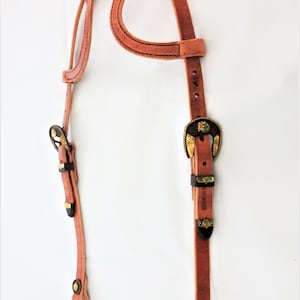 Puede incluir: Un cabezal de cuero marrón para caballos con dos hebillas de latón y detalles decorativos. El cabezal tiene un diseño único con un lazo en la parte superior.