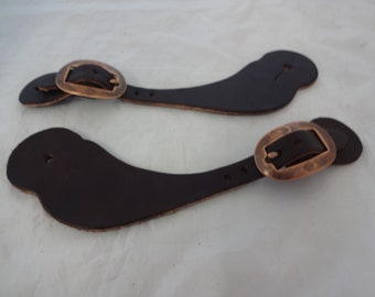Può includere: Un paio di cinturini per speroni in pelle marrone con fibbie in ottone anticato. I cinturini sono progettati per essere fissati a uno sperone e poi a uno stivale.