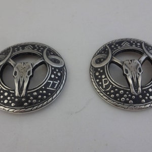 May include: Two silver-toned metal round pendants with a bull skull design. Each pendant has a circle with a bull skull inside, surrounded by a circle with a pattern of dots and a triangle at the top. The pendants have the letters "P" and "II" on the bottom.