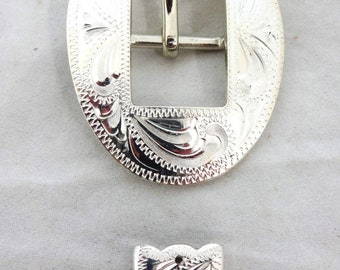 May include: Silver-toned metal belt buckle with an oval shape and intricate engraved floral designs. The buckle has a rectangular opening for a belt strap. A matching silver-toned metal tip is also included.
