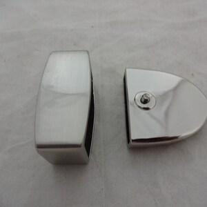 May include: Two silver-toned metal glass clamps for a glass panel. One clamp is rectangular and the other is rounded.