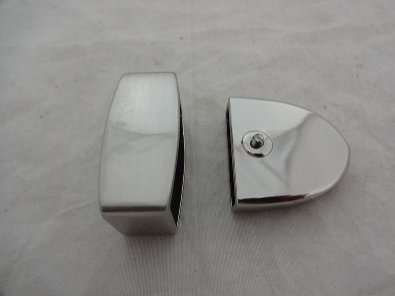 May include: Two silver-toned metal glass clamps for a glass panel. One clamp is rectangular and the other is rounded.