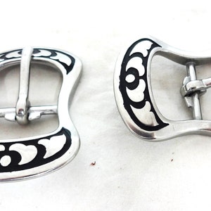 May include: Two silver-toned metal belt buckles with a black floral design. The buckles are designed to be used with a strap.