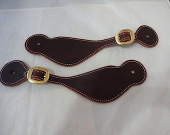 Può includere: Due cinturini per speroni in pelle marrone con cuciture bianche e fibbie dorate. I cinturini sono progettati per essere fissati agli stivali da cowboy.