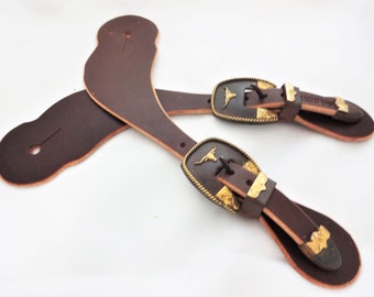 Può includere: Un paio di speroni in pelle marrone con fibbie decorative color oro. Le fibbie presentano un design a testa di bue. Gli speroni sono progettati per l'equitazione e hanno un classico stile western.
