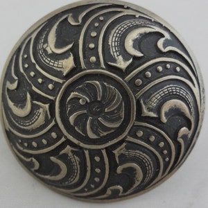 Puede incluir: Un pomo de metal plateado con un diseño intrincado en relieve que presenta patrones en espiral y un motivo circular central. El pomo tiene una forma ligeramente abovedada.