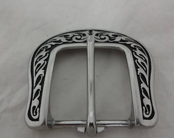 Könnte beinhalten: Silberfarbene Metallschnalle mit einem dekorativen schwarzen Filigranmuster an den Rändern. Die Schnalle hat eine klassische Hufeisenform mit einem einzelnen Dorn zum Befestigen des Gürtels.