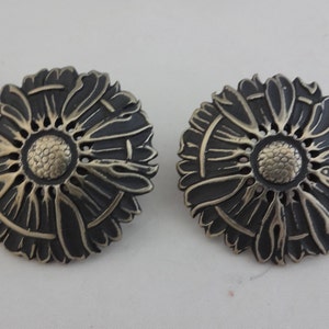 May include: A pair of silver tone earrings with a black enamel flower design. The earrings are round and have a raised center with a textured design.
