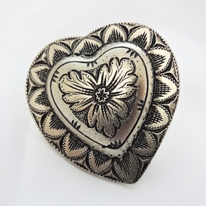 Puede incluir: Un colgante de plata en forma de corazón con un diseño floral. El colgante tiene un diseño detallado e intrincado con una flor en relieve en el centro.