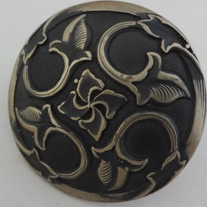 Puede incluir: Un pomo redondo de metal plateado con fondo negro y un intrincado diseño floral.
