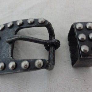 May include: A silver metal belt buckle with a studded design. The buckle is rectangular with a rounded edge and has a silver metal tongue. The buckle is attached to a matching silver metal belt loop.