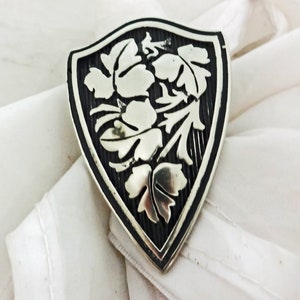 May include: A silver-toned metal brooch in the shape of a shield with a floral design. The shield is black with a silver floral pattern.