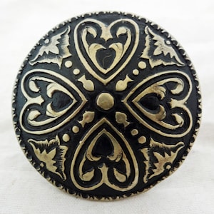 Puede incluir: Un botón redondo de metal negro con un diseño plateado de cuatro corazones estilizados y detalles florales.