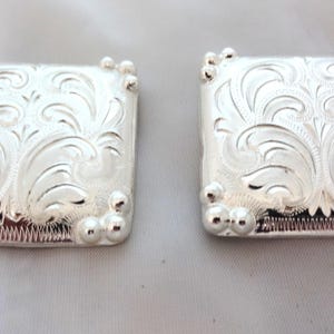 Puede incluir: Dos hebillas de cinturón cuadradas de color plateado con intrincados diseños florales. Cada hebilla tiene bordes redondeados y pequeños detalles de bolas decorativas. Las hebillas probablemente son de metal.