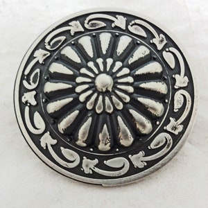 Puede incluir: Un botón de metal plateado con un diseño circular en relieve. El centro del botón presenta una flor estilizada con un centro en relieve. El borde exterior del botón está decorado con un patrón repetitivo de hojas estilizadas.
