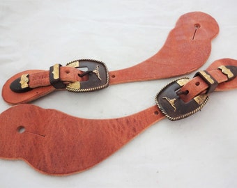 Può includere: Un paio di cinturini per speroni in pelle marrone con fibbie in metallo dorato e marrone. Ogni fibbia presenta un design in rilievo di una testa di toro.