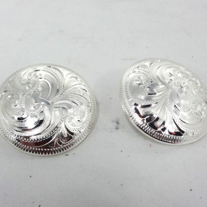 May include: Two silver-colored, round conchos with intricate floral scrollwork. The conchos have a raised, detailed design and a polished finish. These decorative pieces are likely for Western wear.