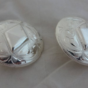 May include: Two silver-toned metal conchos with a diamond-shaped design in the center and intricate floral patterns around the edges.