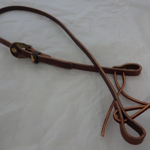 Puede incluir: Una correa de cuero marrón con una hebilla de metal y dos bucles en el extremo. La correa está diseñada para sujetarse a una silla de montar u otro equipo.