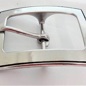 May include: A silver belt buckle with a black border. The buckle has a curved shape and a simple design.