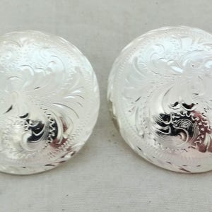 May include: Two silver-colored, round conchos with an engraved floral design. The conchos have a domed shape and a bright, reflective surface. They are likely used for decorative purposes on leather goods.