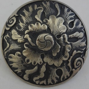 Puede incluir: Un objeto redondo de metal plateado con un intrincado diseño floral. El patrón floral en relieve se establece sobre un fondo oscuro, creando un contraste. El diseño presenta una espiral central y hojas.